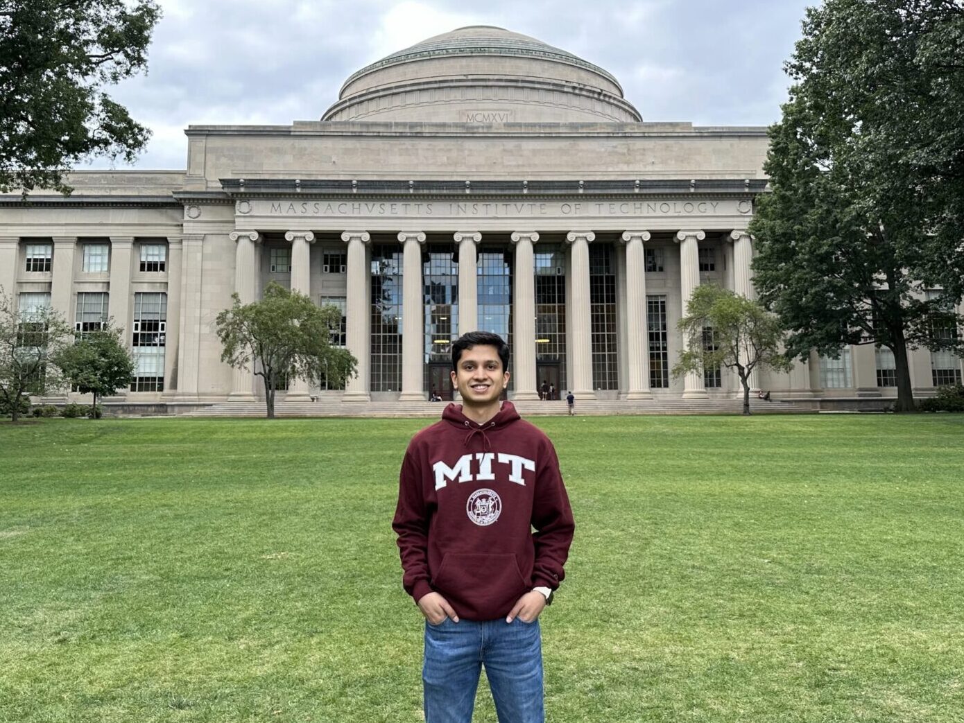 Rishabh Shanbhag pictured in front of the iconic Great Dome building of the Massachusetts Institute of Technology (MIT), wearing a maroon MIT hoodie and smiling.